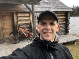 Kregg Ochitwa, physiotherapist, taking a selfie in front of his cottage with a bike also in the background.